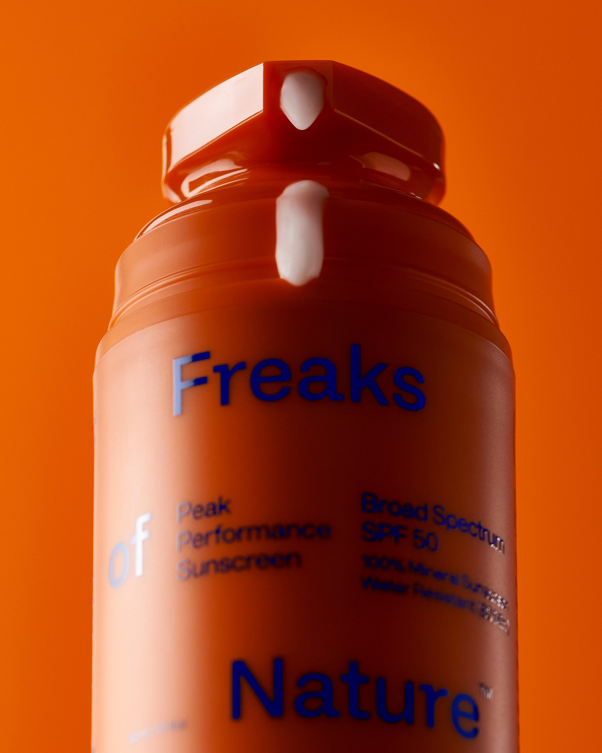 Close-up of an orange bottle of Freaks of Nature Skincare sunscreen against an orange background. The label reads "Peak Performance SPF50, Broad Spectrum SPF 50, 100% Mineral Sunscreen with vegan spider silk protein, Water Resistant 80 min." The bottle has a white pump dispenser on top.