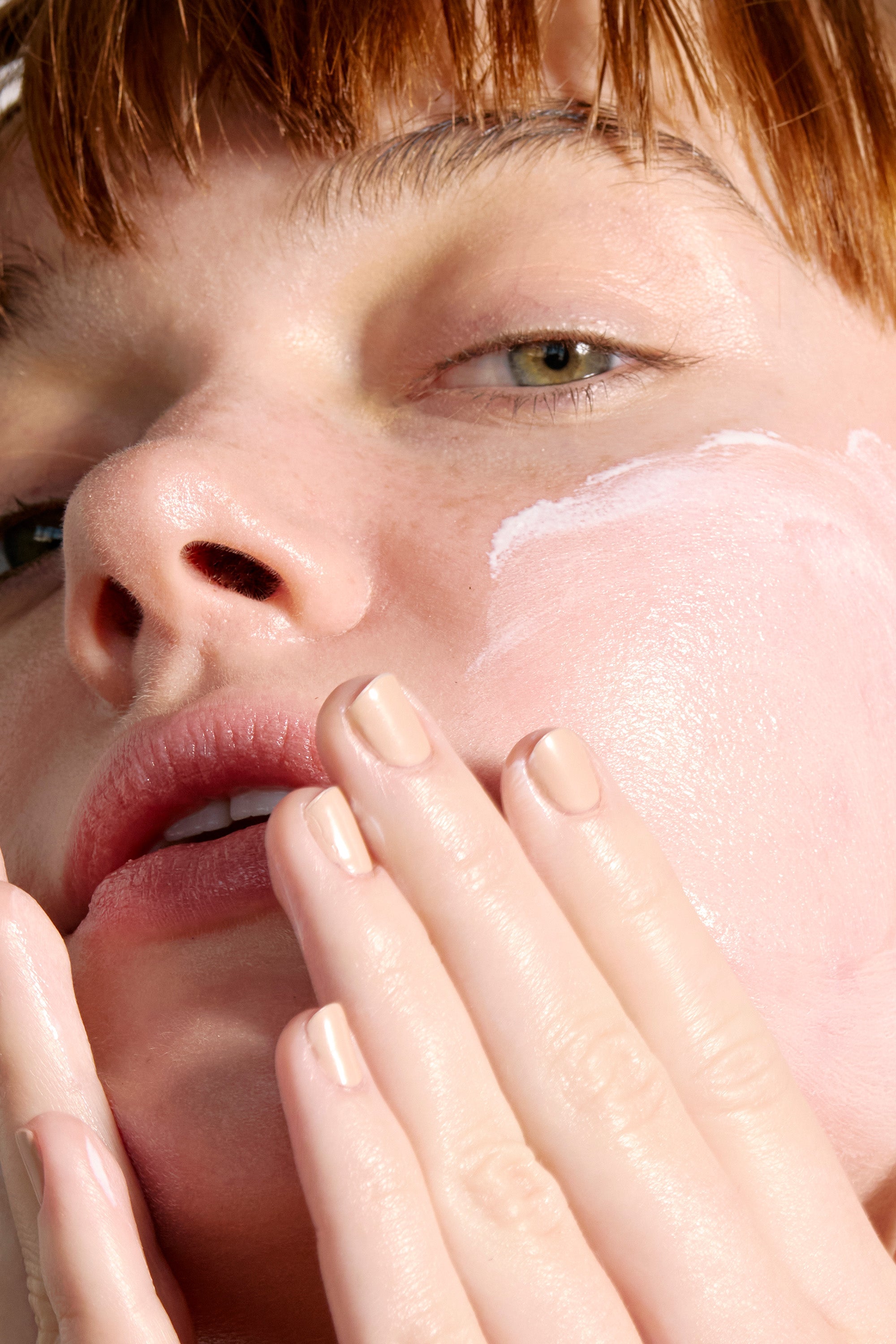 Close-up of a person applying Peak Performance SPF50 - SAMPLE by Freaks of Nature Skincare, a creamy pink product infused with vegan spider silk protein, to their cheek. They have short, light-colored hair and light eyes, and their nails are neatly manicured with a neutral polish. The skincare product is being gently rubbed into the skin with their fingers.