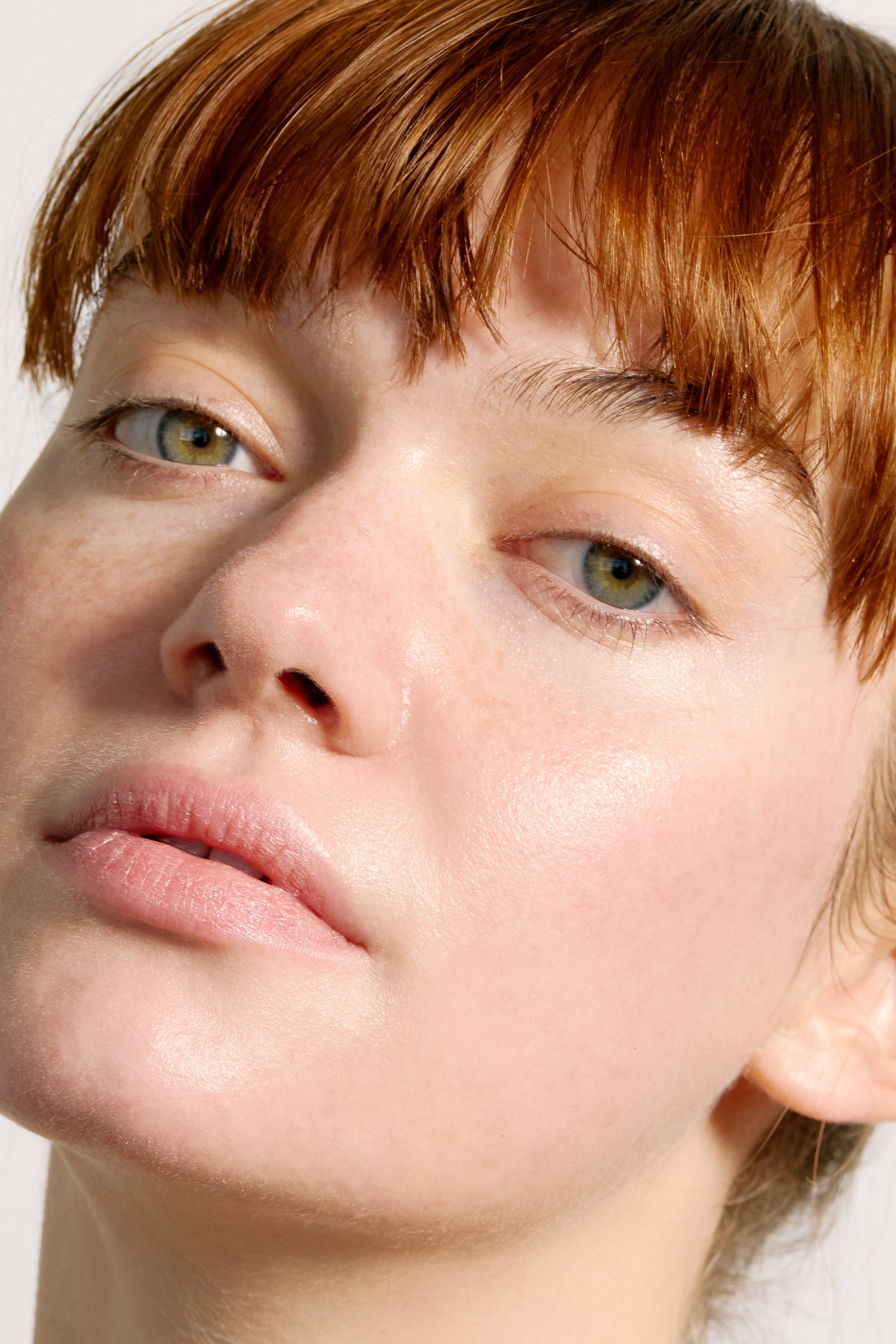 A close-up of a person with fair skin and red hair with bangs looking slightly off camera. Their bare face showcases clear skin with a natural look, highlighting the effects of Freaks of Nature Skincare's Peak Performance SPF50 - SAMPLE. The neutral background keeps the focus on their face, enhanced by the nourishing benefits of red algae MAA extracts.