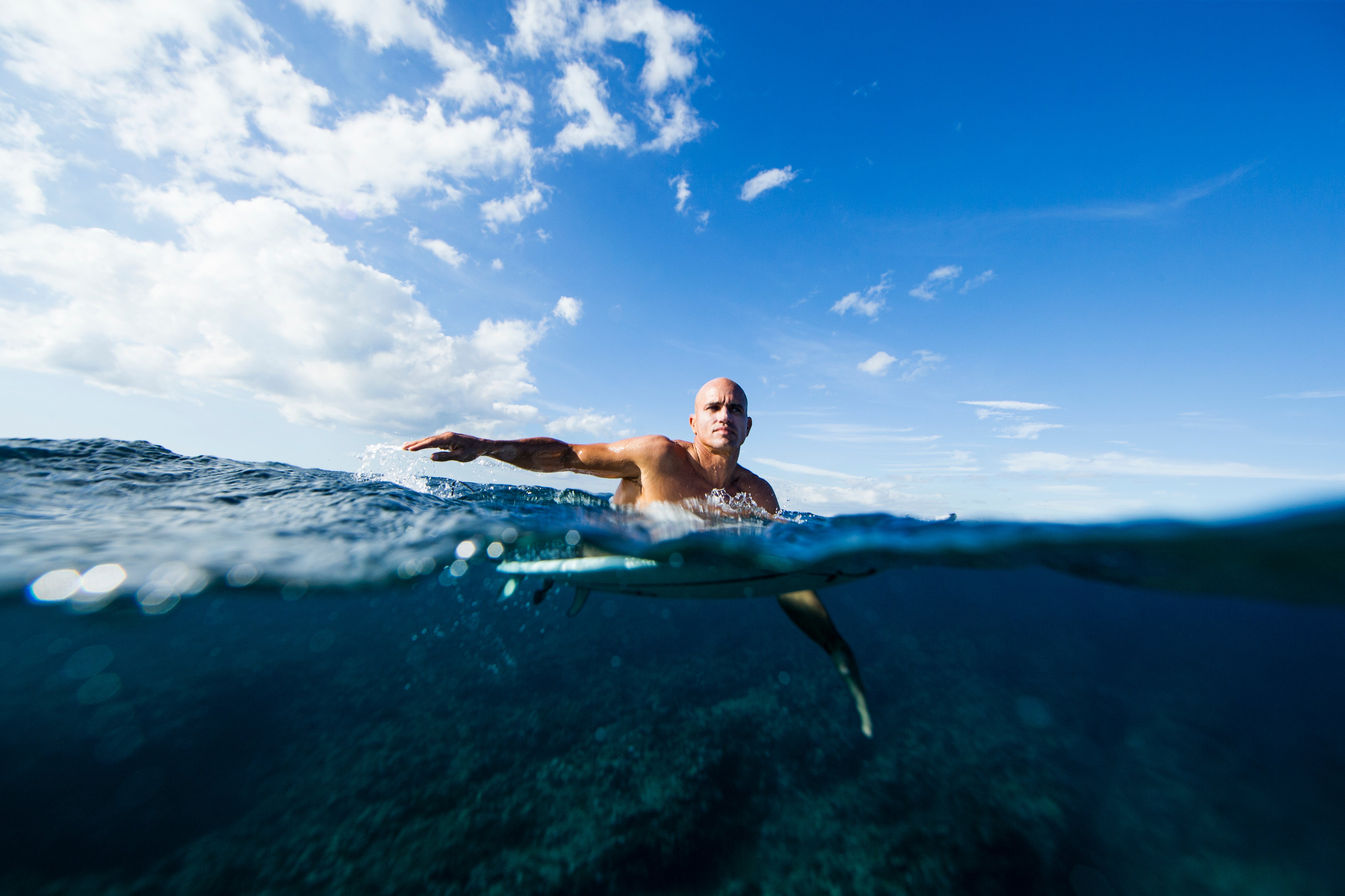 A man paddles on a surfboard in clear blue ocean water under a partly cloudy sky, protected by Freak Trio from Freaks of Nature Skincare. The image is captured from a water-level perspective, showing half above and half underwater, with the man in the center of the frame.