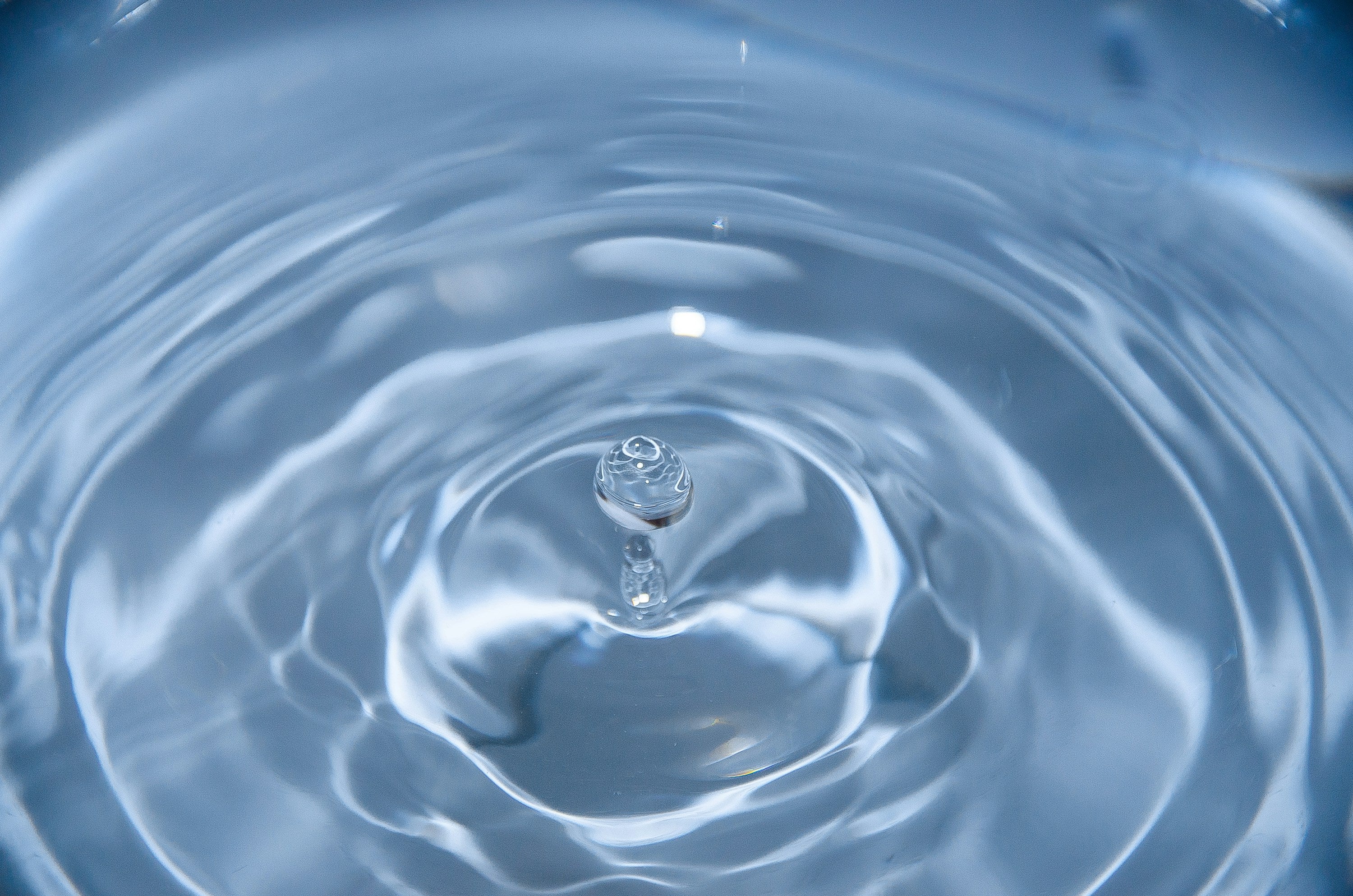 Close-up image of a single water drop splashing into a body of water, creating ripples in concentric circles around the impact point. The drop and surrounding water are clear and translucent, with a blue hue due to the lighting.