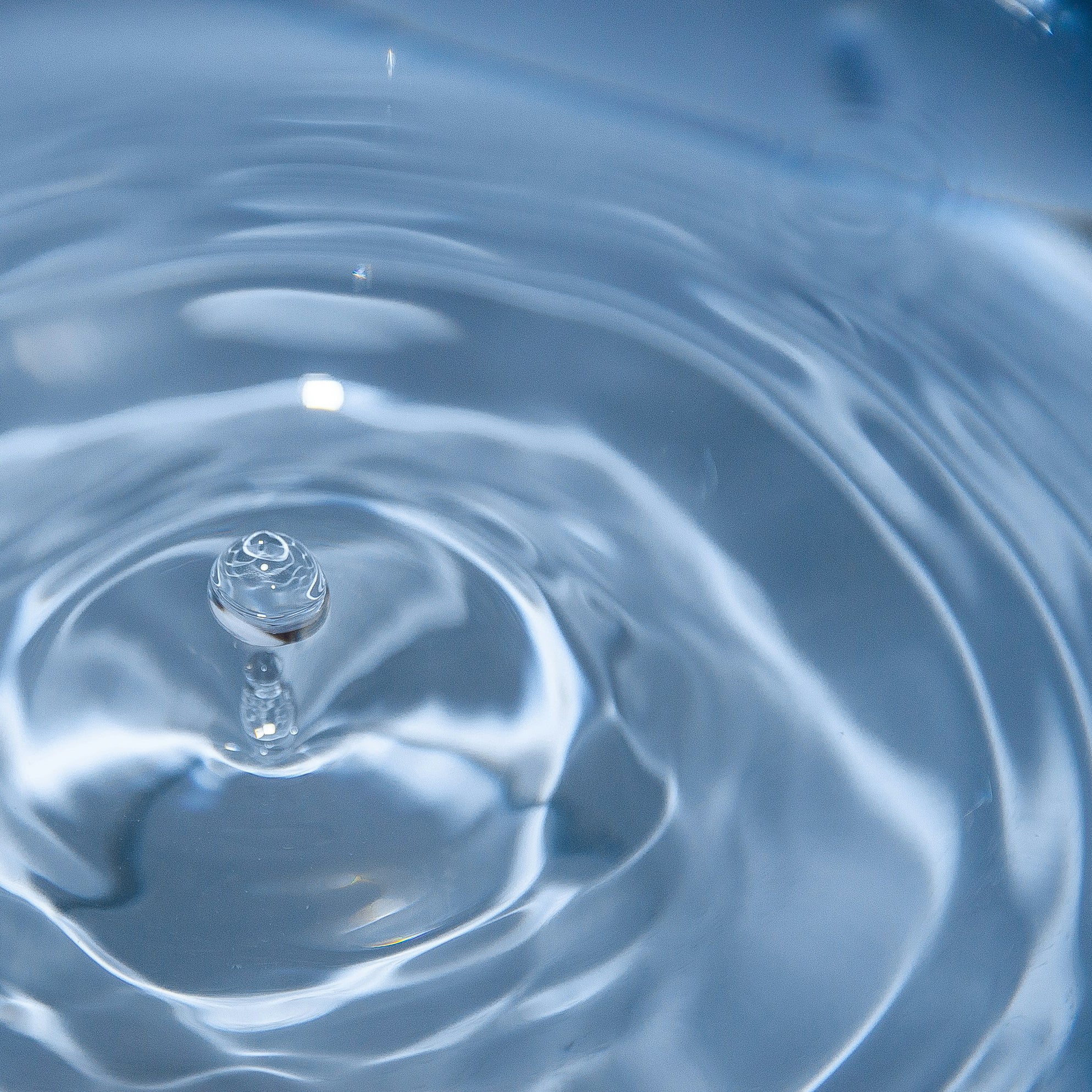 A close-up shot of a droplet of water creating ripples as it splashes into a calm water surface. The image captures the moment the droplet makes contact, forming concentric circles spreading outward in the clear blue water.
