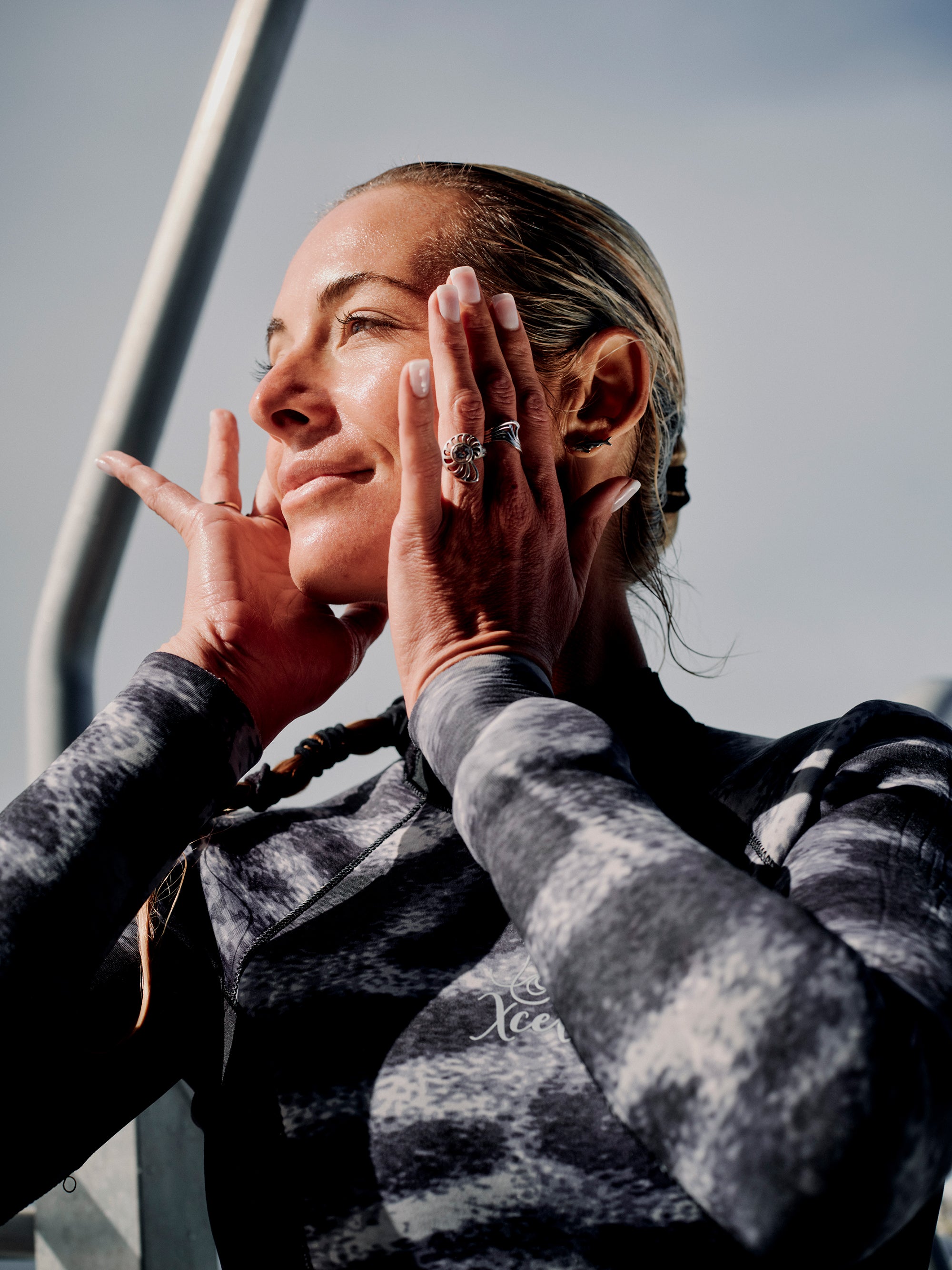 A woman in a wetsuit touches her face with both hands, looking upwards with a serene expression. She has her hair pulled back and is wearing several rings on her fingers. Perhaps it's the Deeper Dive Moisturizer by Freaks of Nature Skincare enriched with vegan shark Squalane that adds to her glow. The background features railings and a clear sky.