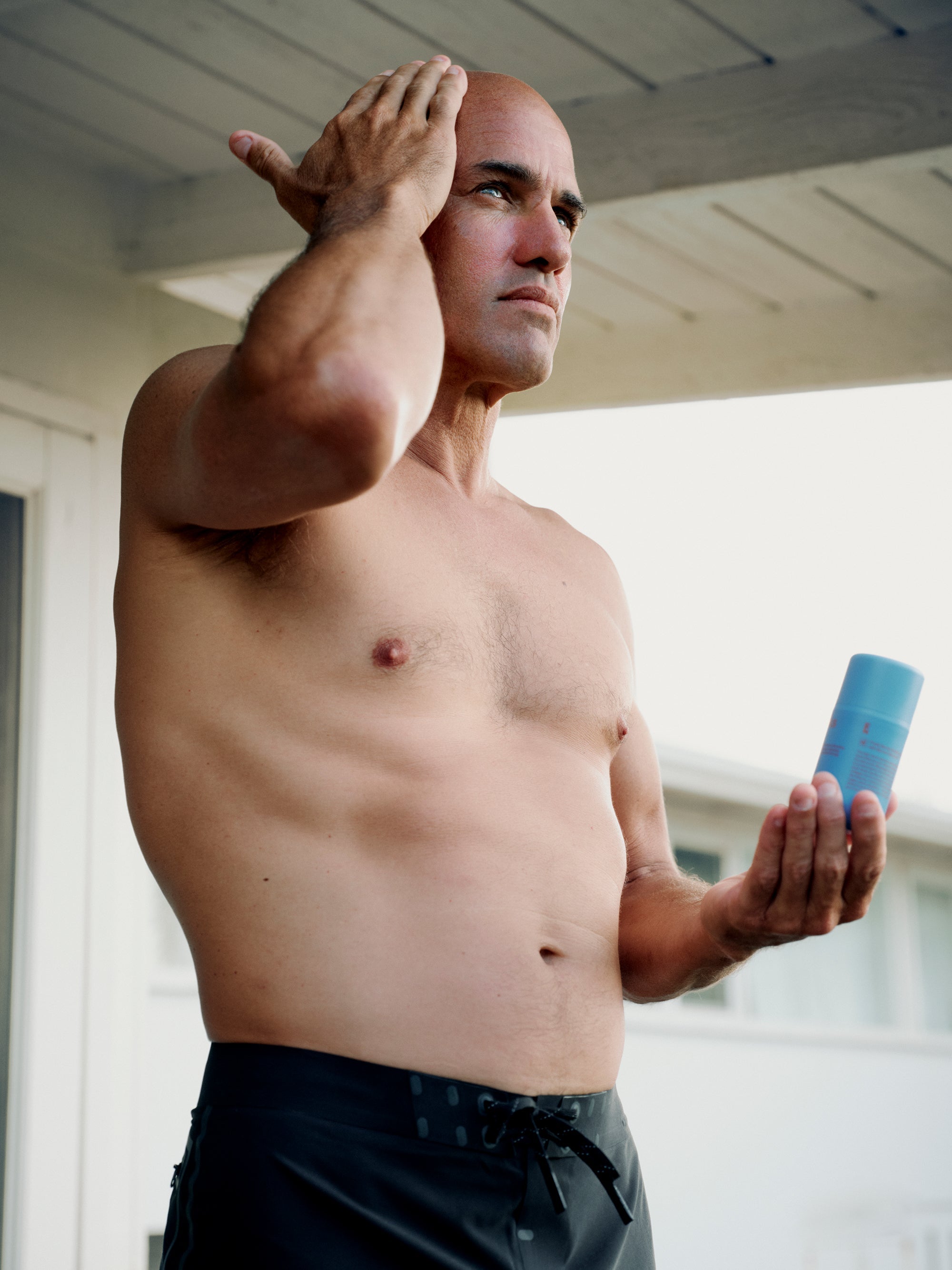 A shirtless man with a serious expression stands under a wooden roof, applying a product to his face with his right hand while holding a blue container in his left hand. He has short hair and wears dark shorts. In the background, there is a white building.