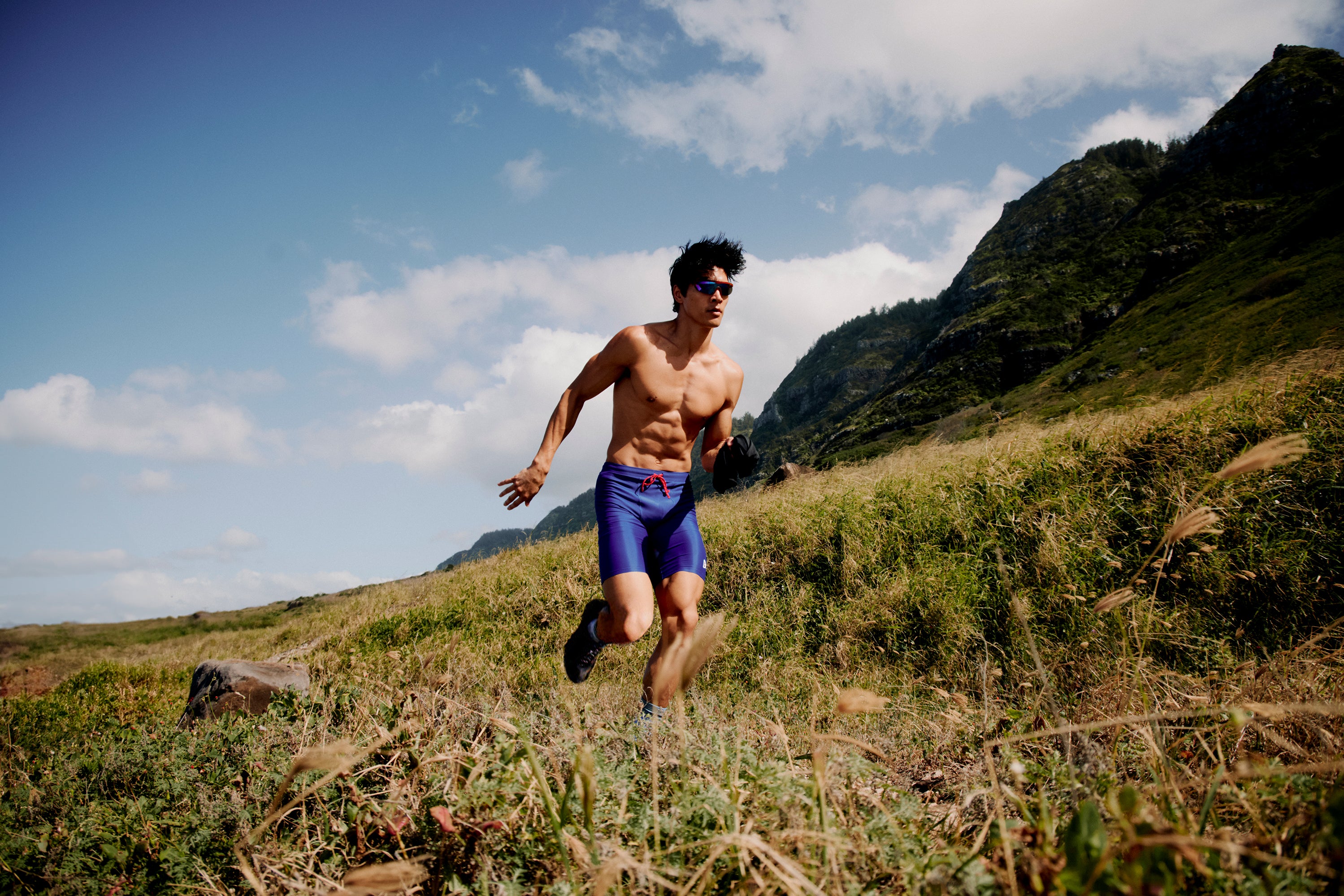 A shirtless person with short black hair runs through a grassy landscape under a clear blue sky. They are wearing blue shorts and sunglasses. Steep hills covered in vegetation are visible in the background.