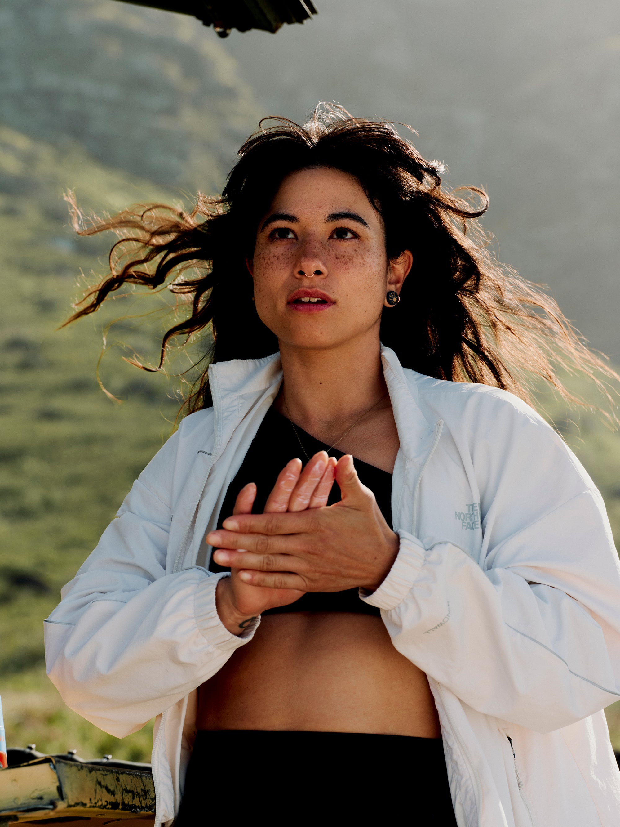 A person stands outdoors with a mountainous background, wearing a white jacket over a black top. Their long hair is flowing in the wind, and they are looking up slightly while clasping their hands together, enjoying the bright and sunny day. They protect their skin with Daily System from Freaks of Nature Skincare for lasting freshness.