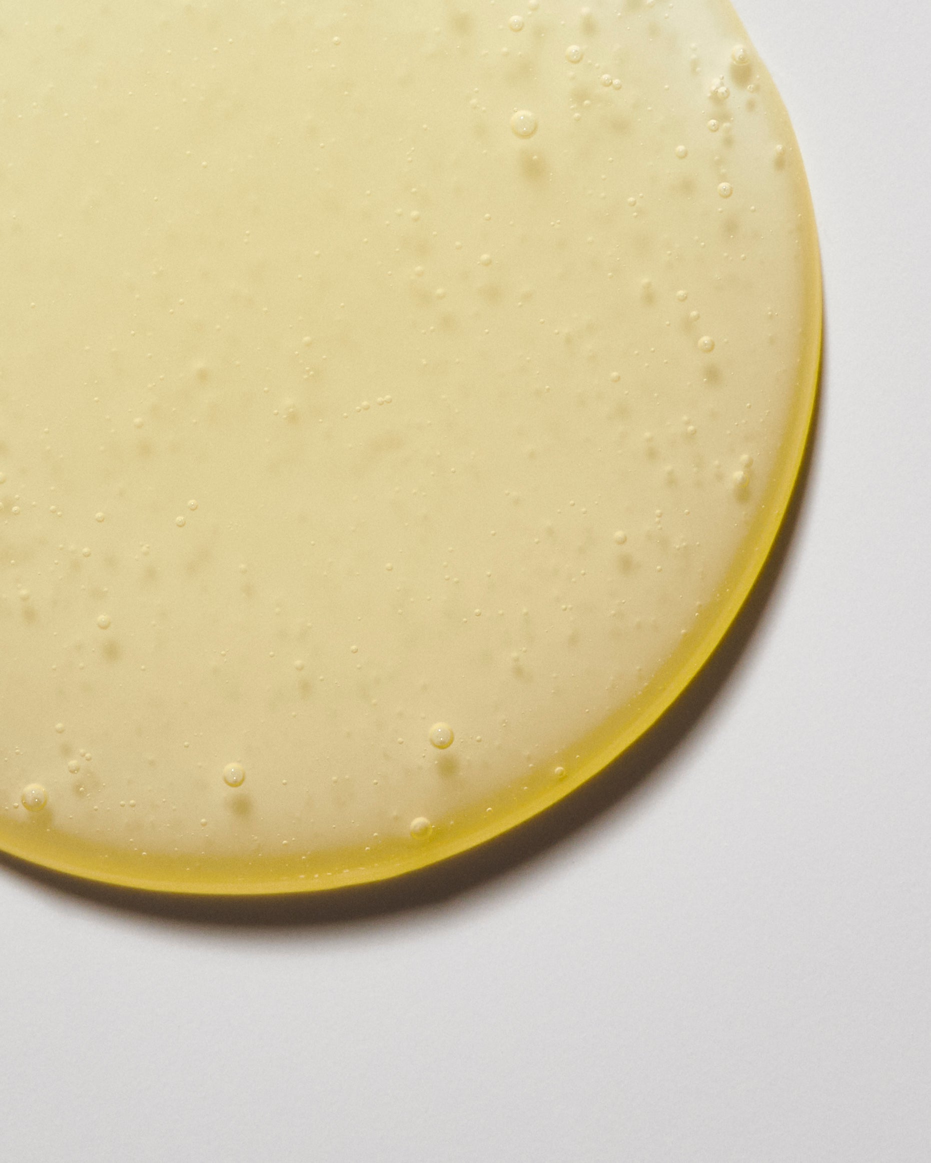 A circular puddle of pale yellow liquid, with a slightly glossy surface and small air bubbles scattered throughout, resembling Deeper Dive Moisturizer by Freaks of Nature Skincare, rests on a white background.
