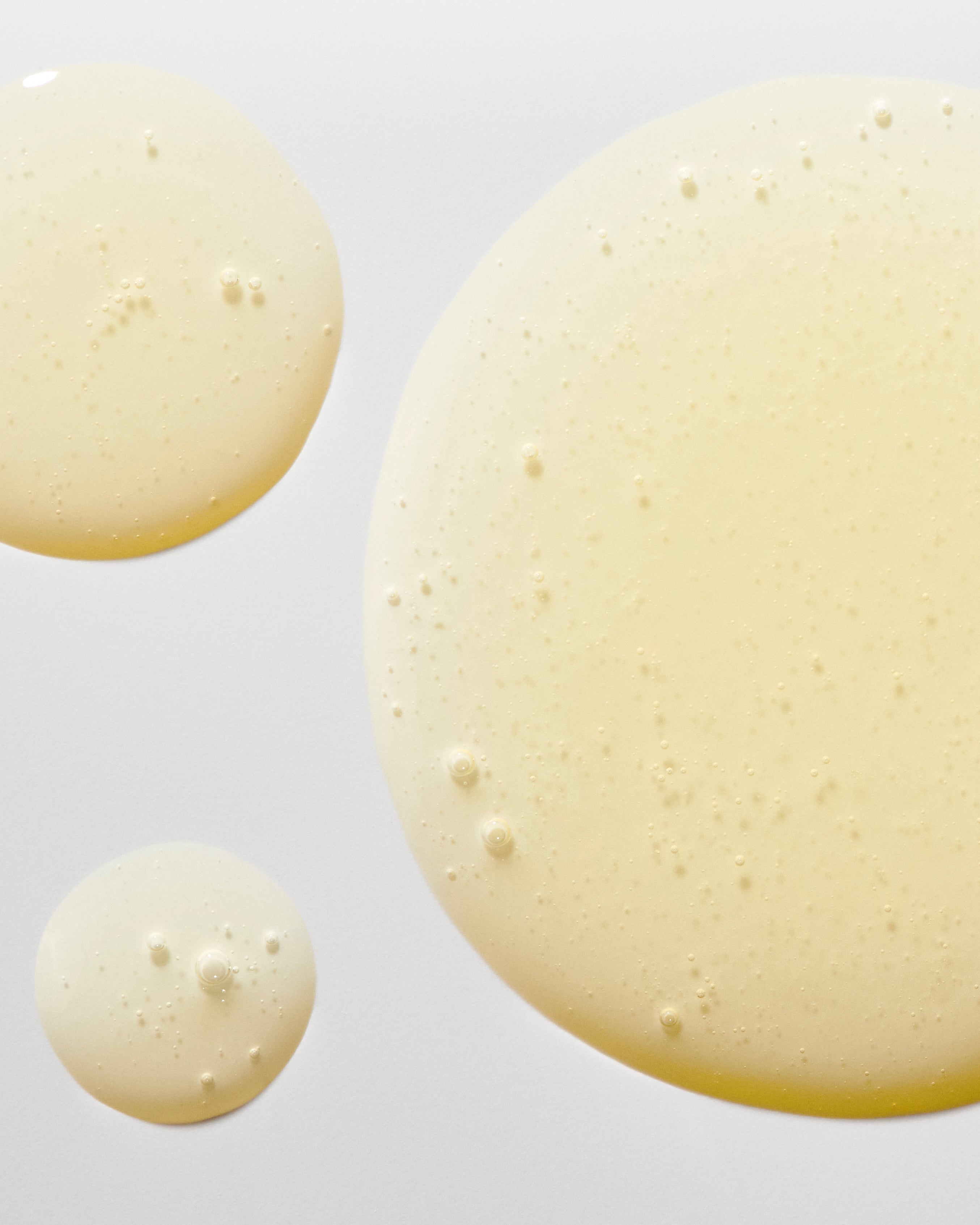 Close-up image of three translucent, amber-colored drops of Freaks of Nature Skincare Daily System on a light background. The slightly viscous drops, enriched with SPF30 Sunscreen and vegan Squalane, contain tiny air bubbles that create a subtle, uneven surface.