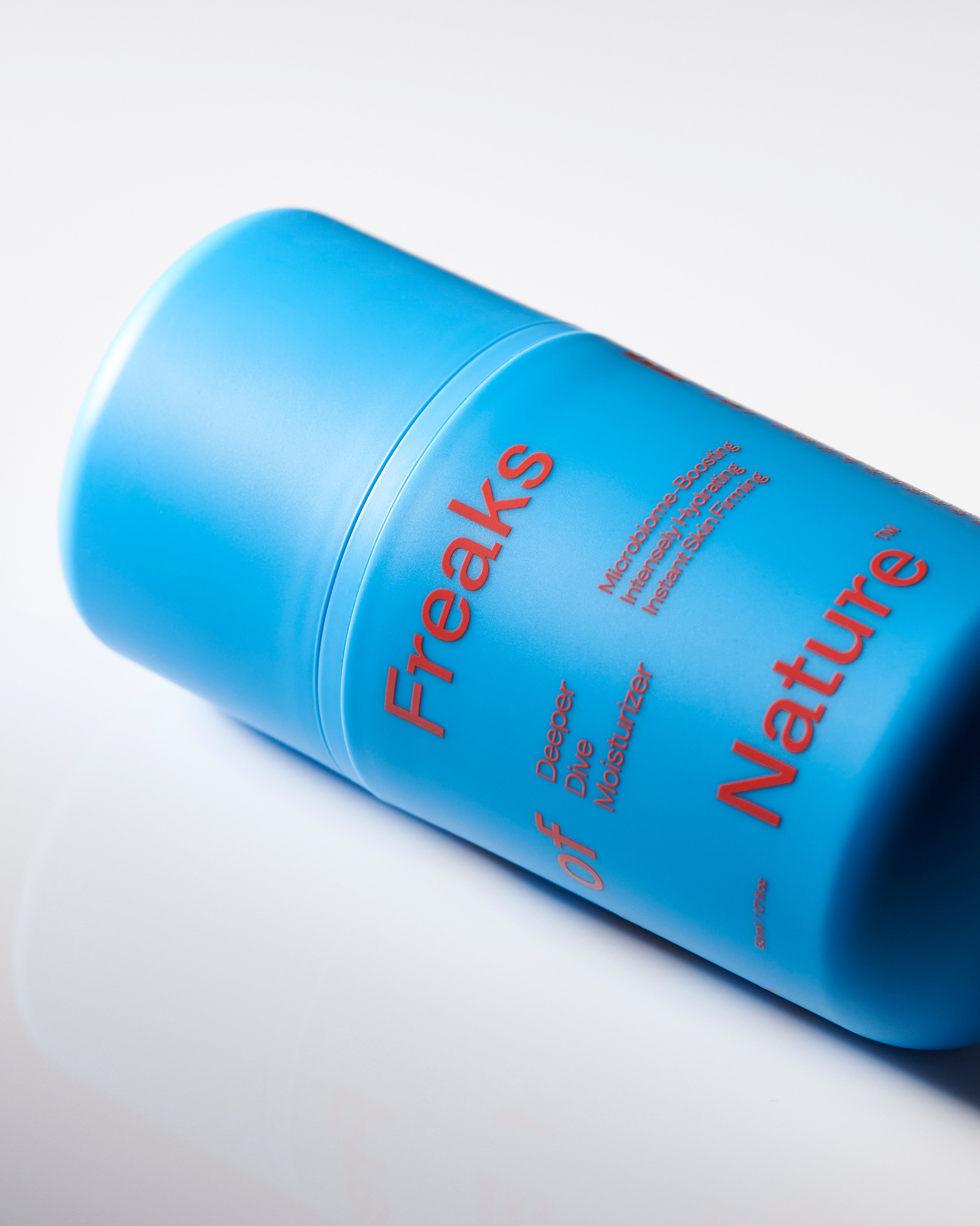 A blue cylindrical container with red text, labeled "Freaks of Nature Skincare," indicating its "Daily System" formula enriched with vegan Squalane. Additional text and product details are visible. The container lies on its side against a plain white background, casting a subtle reflection.