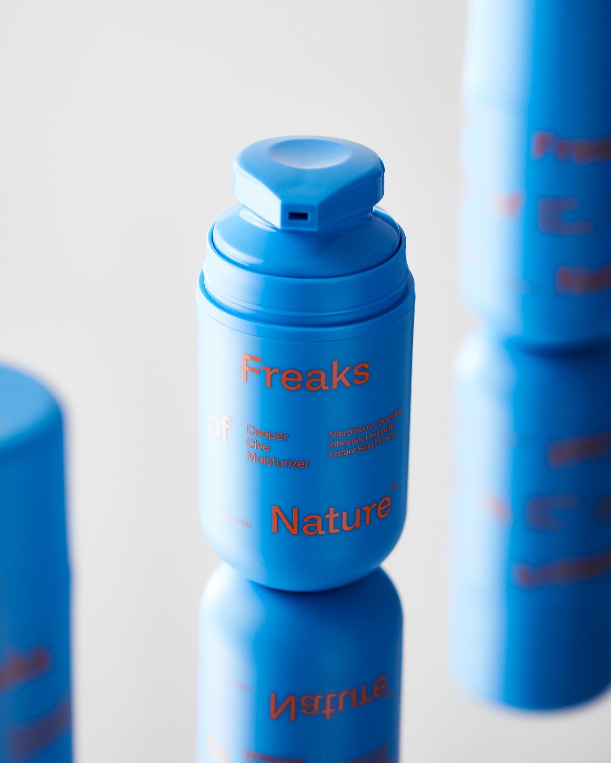 A vibrant blue cylindrical bottle labeled "Freaks of Nature Skincare Deeper Dive Moisturizer" with red text, featuring a flip-top cap. Infused with vegan shark Squalane, it promises optimal hydration and microbiome-balancing. The bottle stands upright on a reflective surface, with partial reflections of similar bottles in the background.