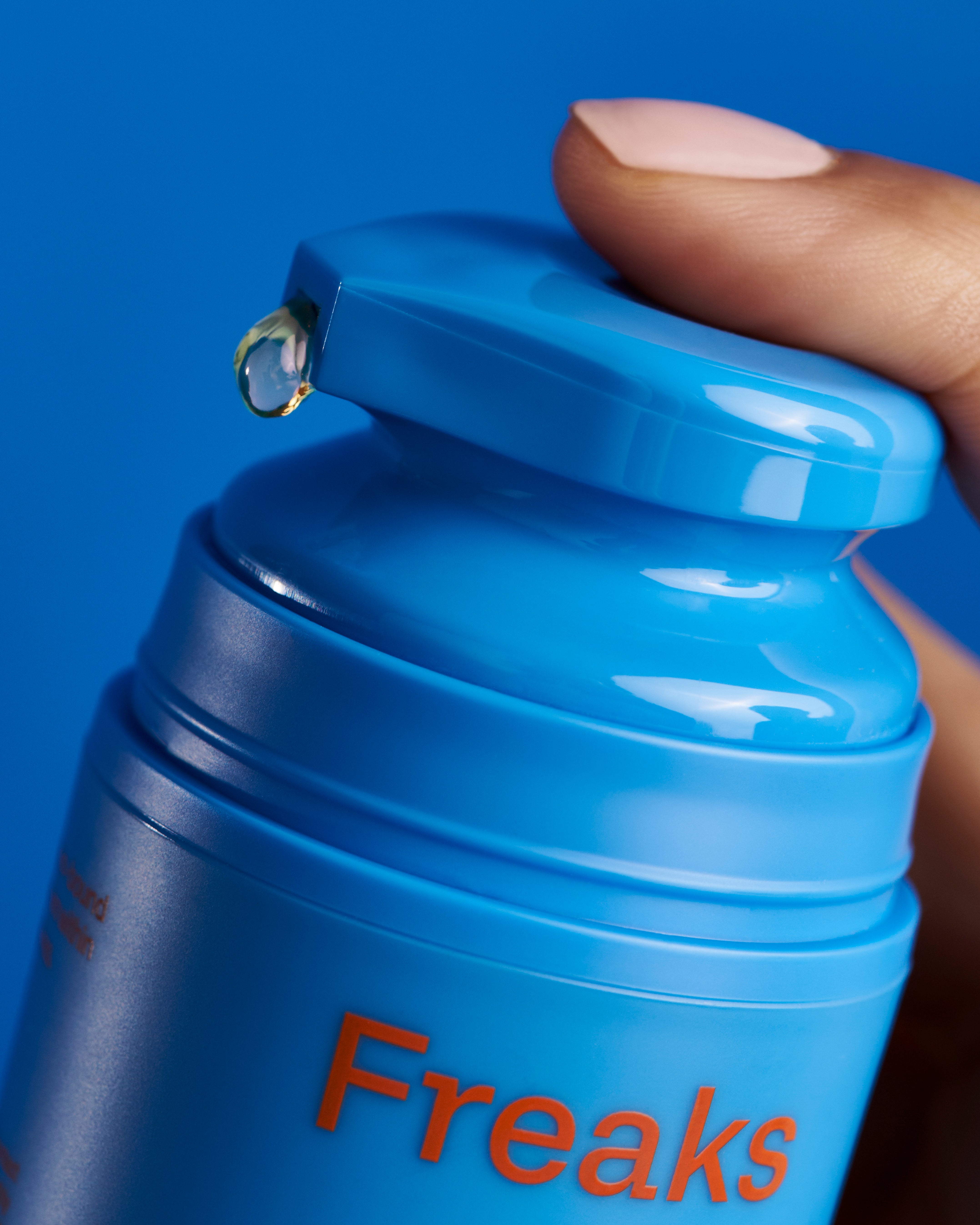 A close-up of a hand holding a blue cylindrical bottle labeled "Freaks of Nature Skincare," which contains Deeper Dive Moisturizer. A clear liquid droplet is visible at the opening of the bottle's cap. The background is a solid blue color, emphasizing its microbiome-balancing moisturizing serum inside.