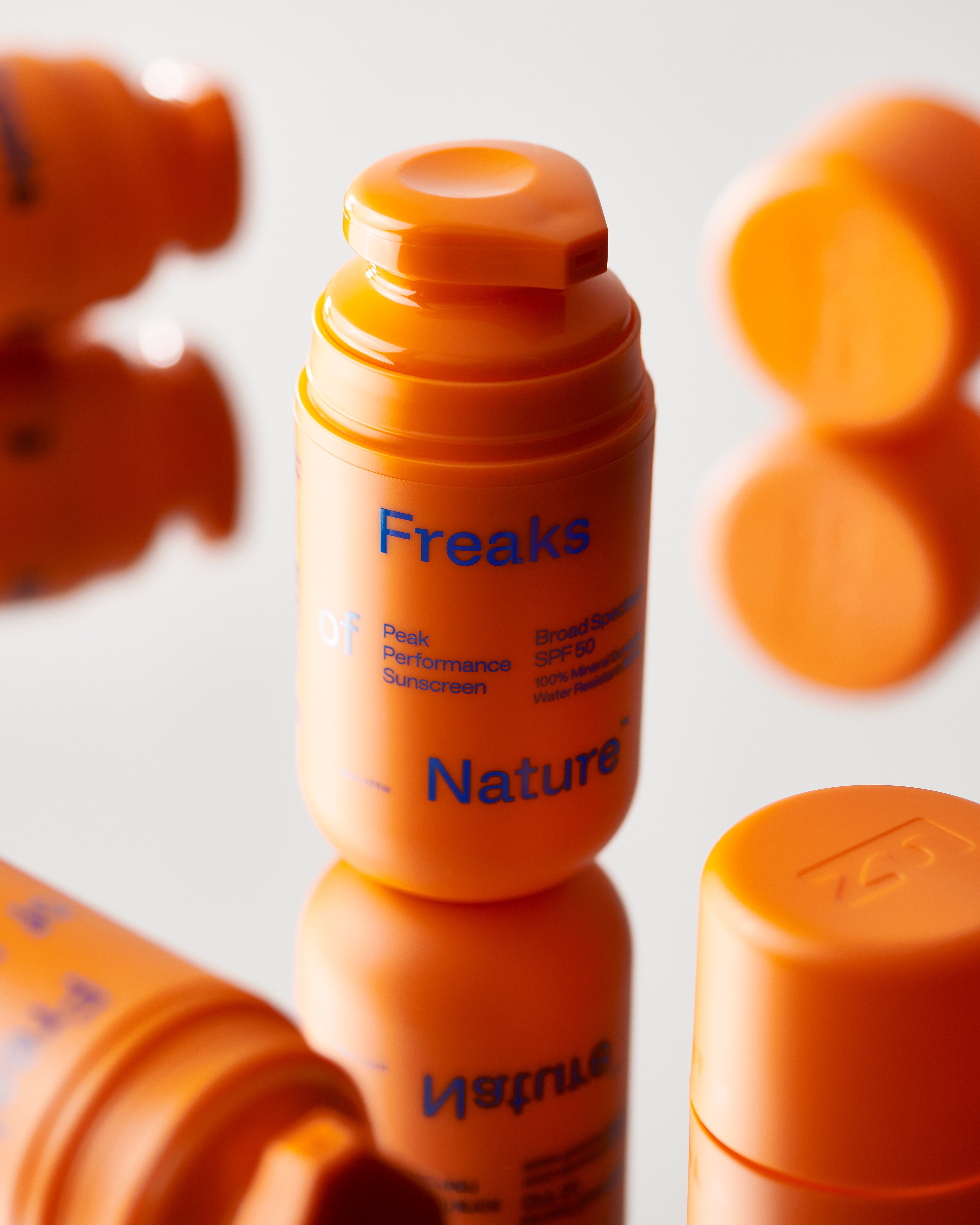 Close-up of several orange cylindrical bottles labeled "Freaks of Nature" containing performance sunscreen with SPF 50. The bottles have flip-top caps and blue text on the label, with some bottles in the foreground and others blurred in the background.