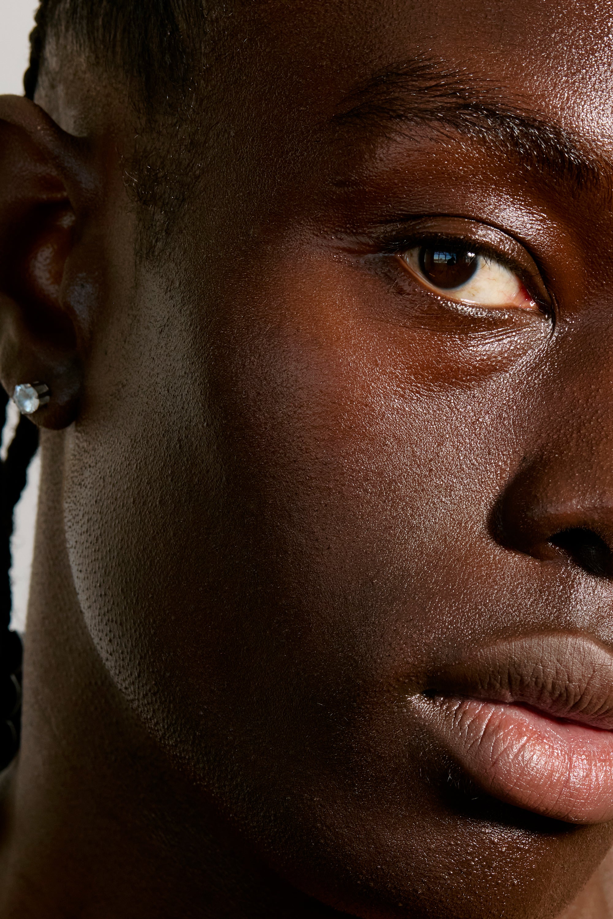 Close-up of half of a person's face with dark brown skin, focusing on the right eye, ear, and part of the nose and lips. The person has braided hair and is wearing a small, diamond stud earring. The image highlights the texture and details of the skin, reflecting Freaks of Nature Skincare's Deeper Dive Moisturizer's nourishing effects.
