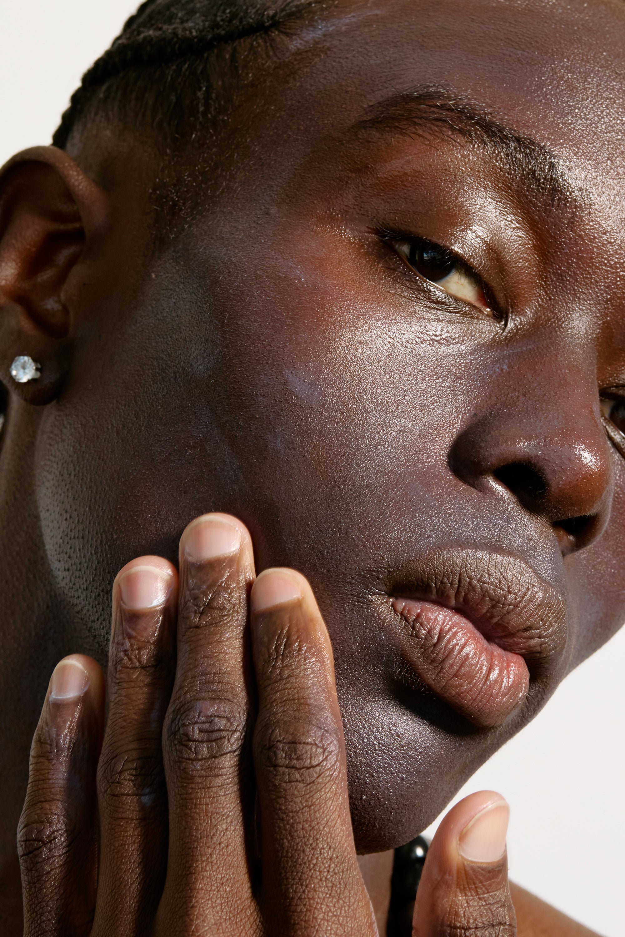 Close-up of a person with dark skin, short braided hair, and a small diamond earring. They are gently touching their cheek with well-groomed nails. The lighting highlights their smooth, dewy complexion thanks to the Freaks of Nature Skincare Daily System and subtle shimmer on their eyelids.