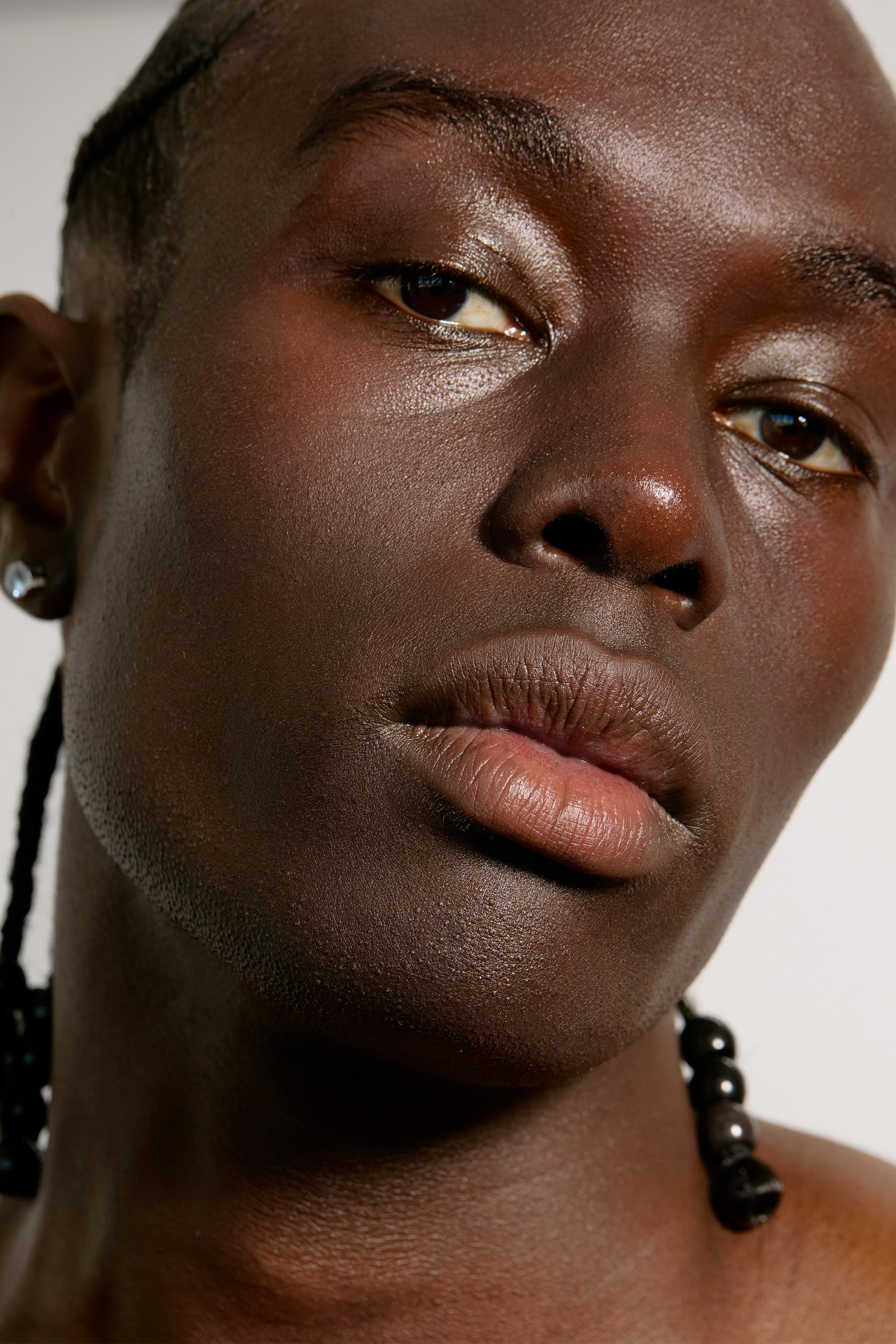 Close-up portrait of a person with dark skin, braided hair adorned with beads, and wearing a small earring. Their head is tilted slightly, reflecting the calm confidence of someone who values self-care. The background is plain white, emphasizing their serene expression and radiant complexion likely nourished by Daily System from Freaks of Nature Skincare.