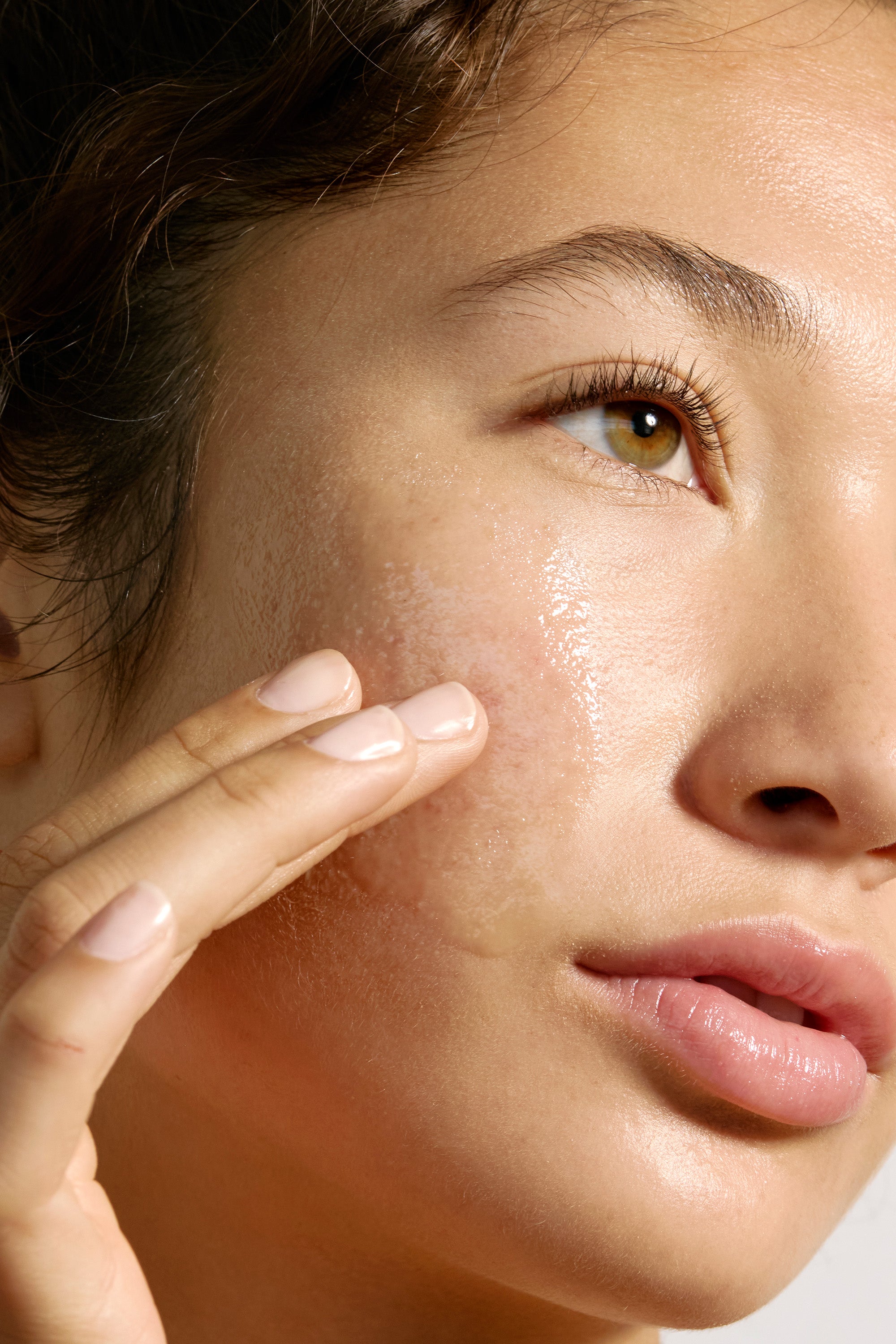 Close-up of a person applying Deeper Dive Moisturizer from Freaks of Nature Skincare to their cheek. The image highlights the person's smooth, dewy skin, focusing on their fingers gently massaging the microbiome-balancing product in a circular motion. Their expression is calm and relaxed.