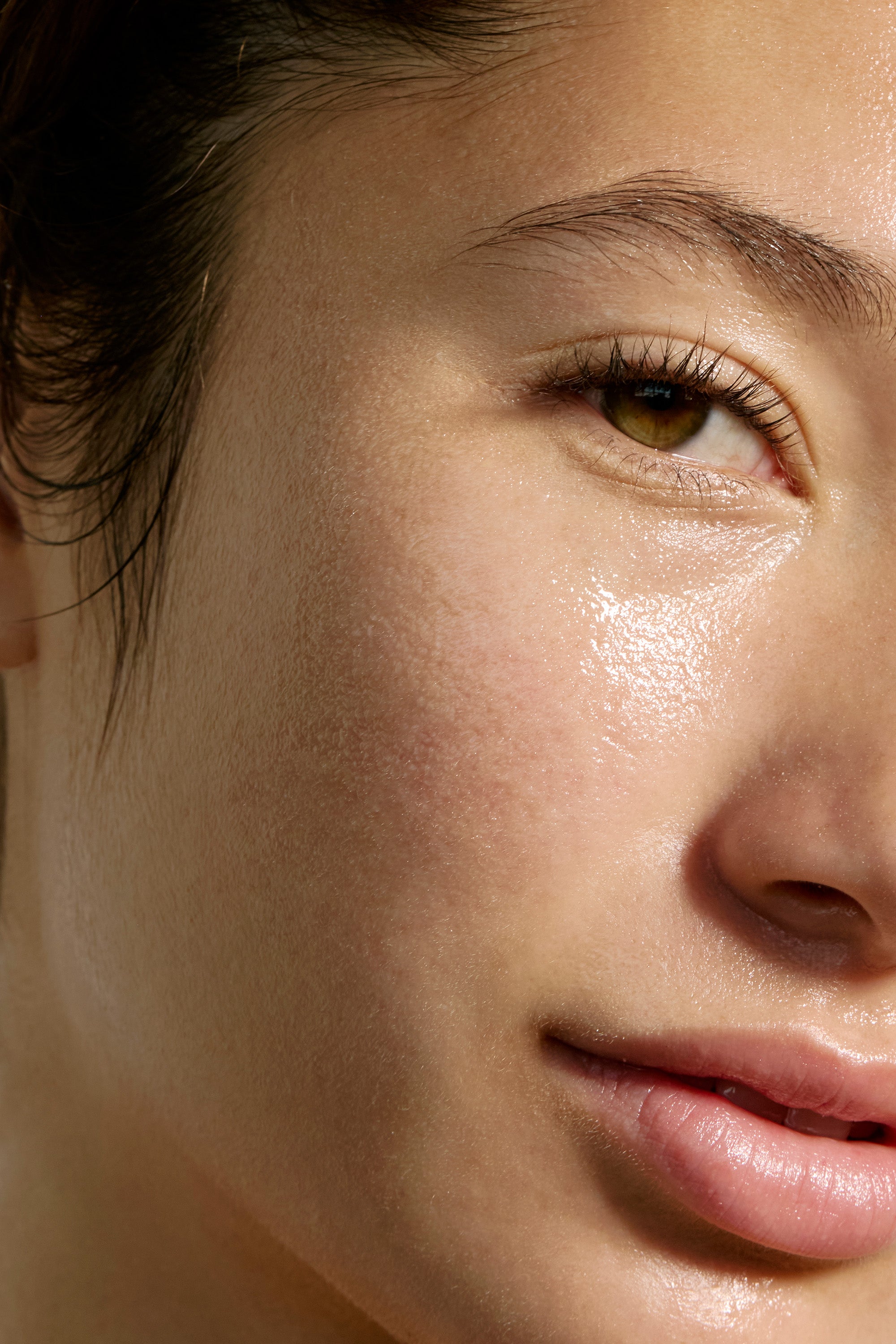 Close-up of a person's face with dewy, radiant skin. The image highlights the cheek, nose, lips, and one eye, showcasing a smooth, natural complexion. Strands of dark hair frame the face. The lighting emphasizes the skin's luminous texture provided by Deeper Dive Moisturizer from Freaks of Nature Skincare.