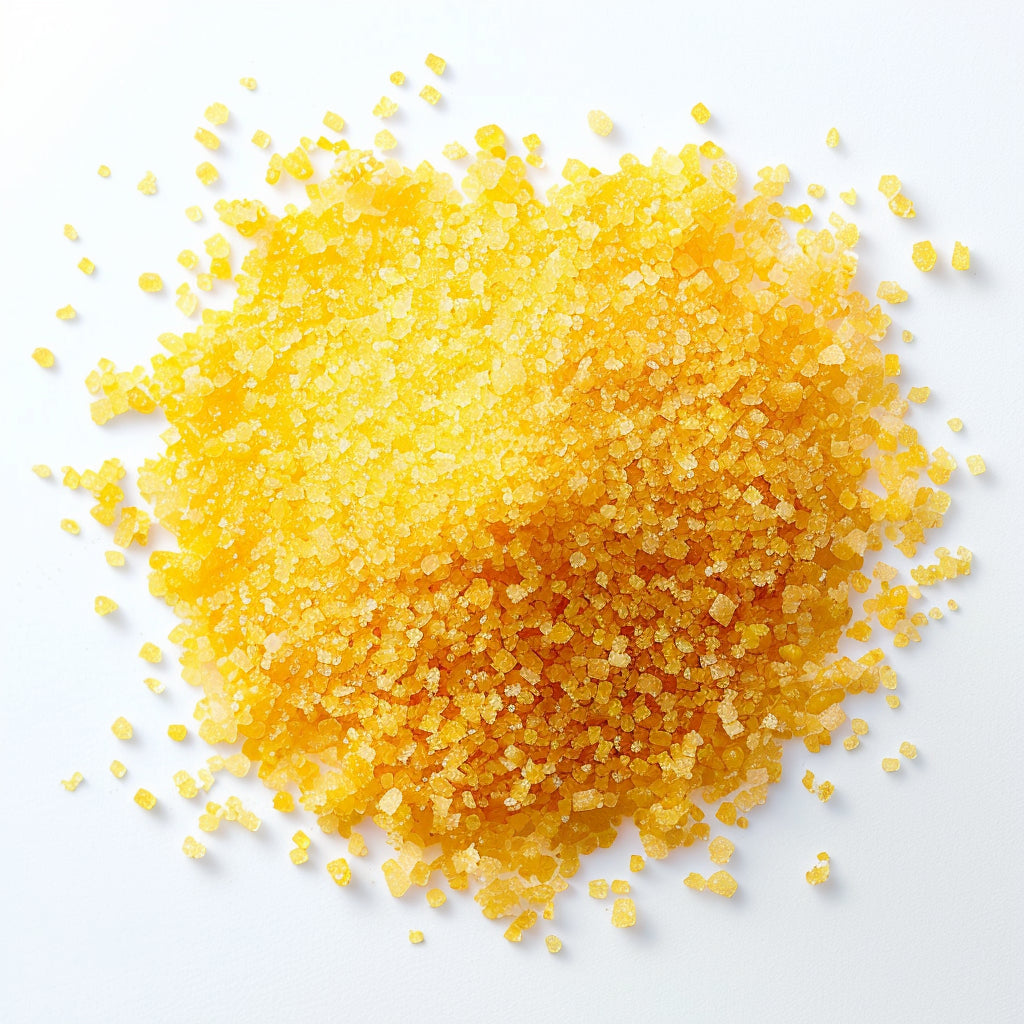 A close-up of bright yellow granulated sugar scattered in a heap on a white surface. The sugar crystals appear coarse and slightly irregular in shape, creating a vibrant, textured pile.