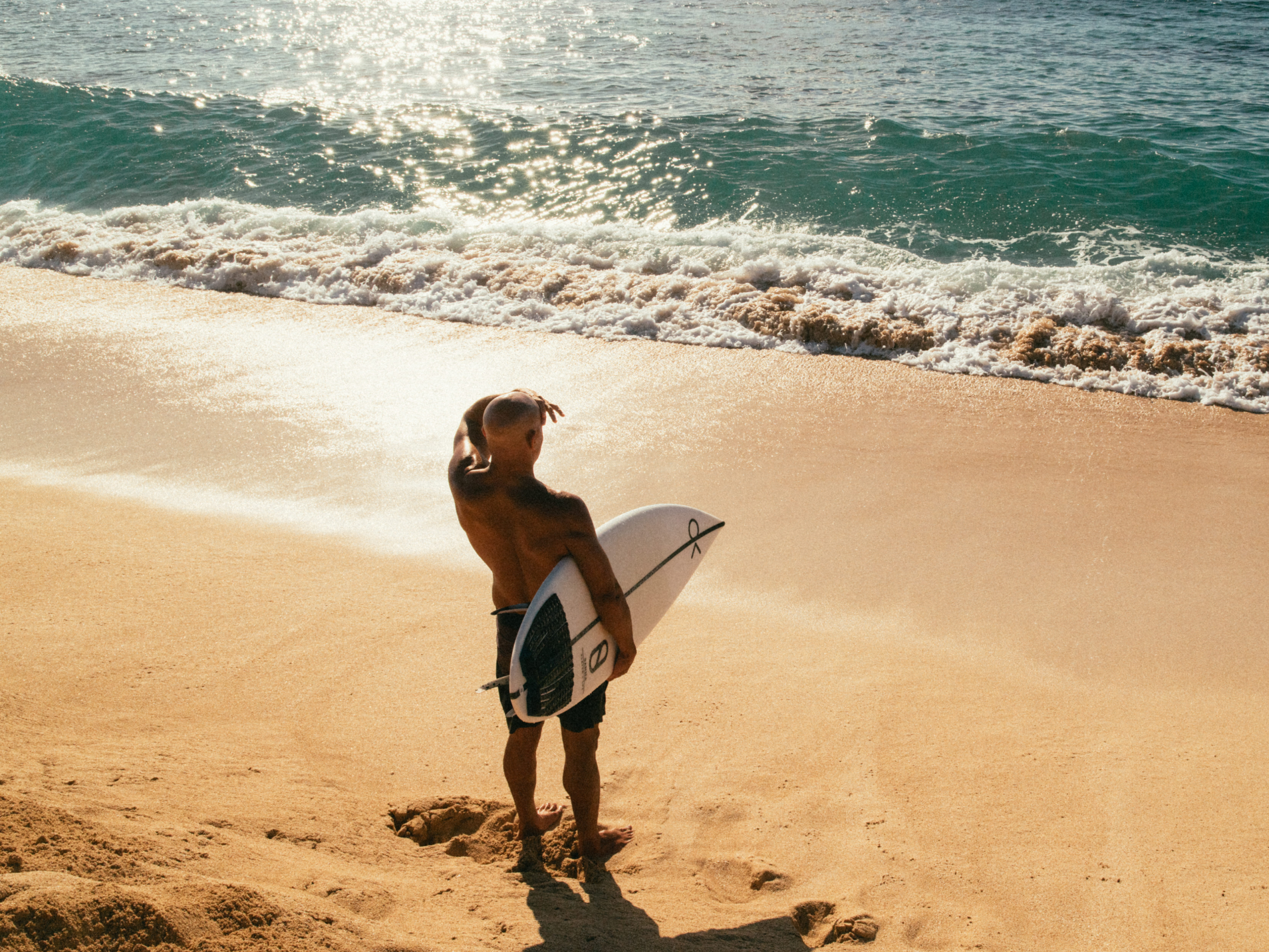 Kelly holding a surfboard on a sandy beach with ocean waves in the background
