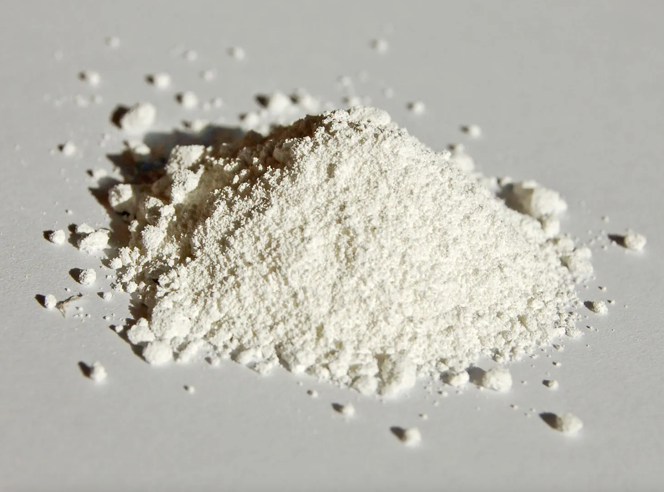 A small pile of white powder on a plain surface. The powder has a fine, grainy texture and is spread slightly around the main pile. The background is a clean, flat surface.