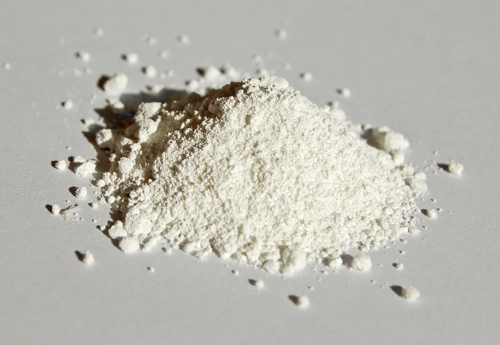 A small mound of fine, white powder is scattered on a light gray surface, with some particles spread loosely around it. The powder appears smooth and slightly clumpy, suggesting a consistency similar to flour or powdered sugar.