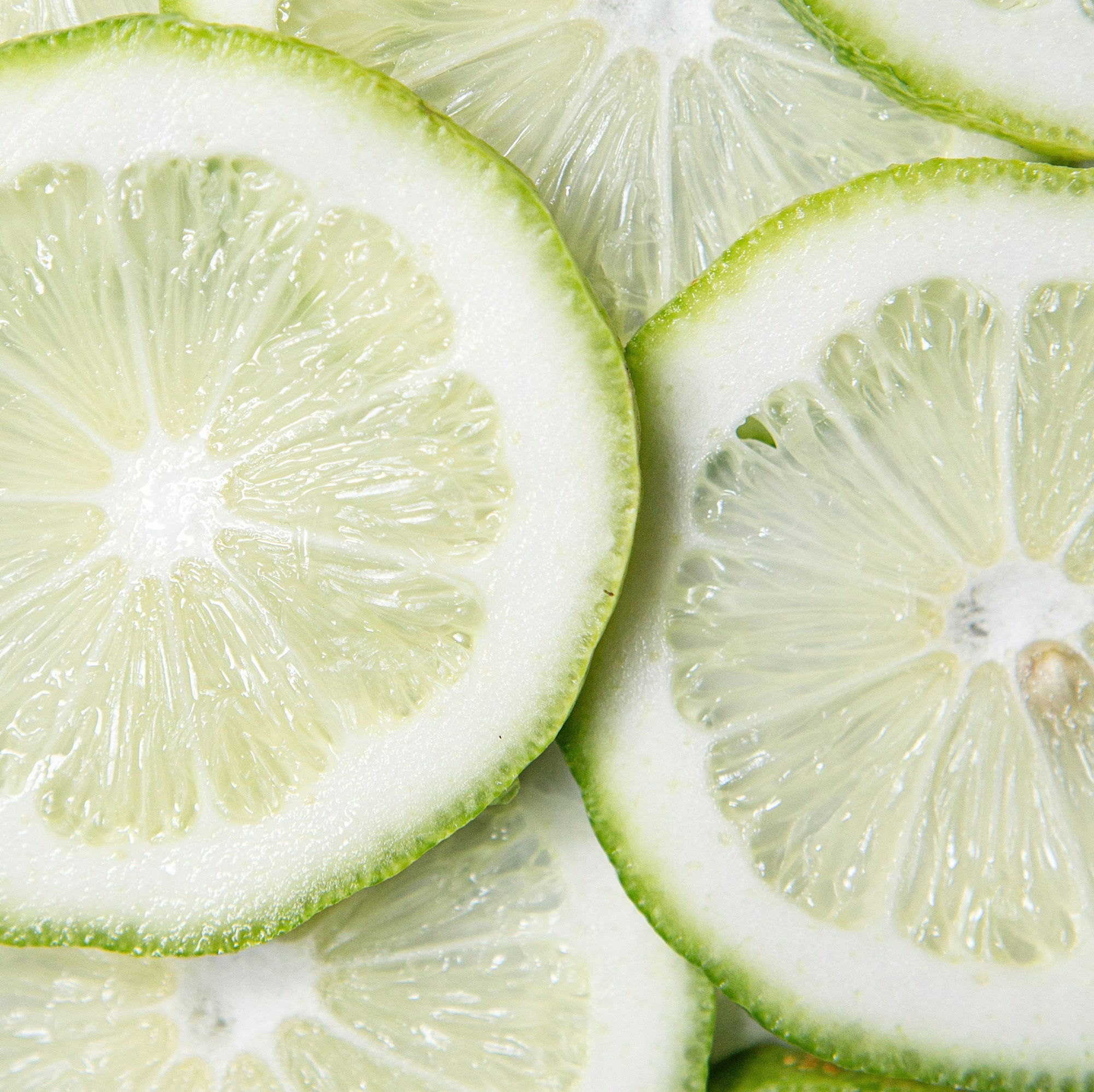 Close-up image of several thinly sliced limes arranged in an overlapping pattern. The slices display a gradient from green outer edges to translucent, pale green centers, showcasing the citrus fruit's texture and juiciness.