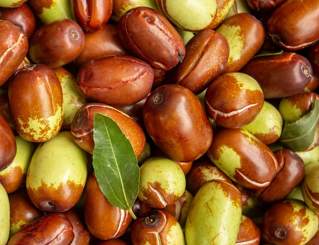 A close-up of a pile of jujube fruits in varying stages of ripeness, with colors ranging from green to reddish-brown. Two green leaves are also visible among the fruits. The surface texture of the fruits appears smooth and glossy.