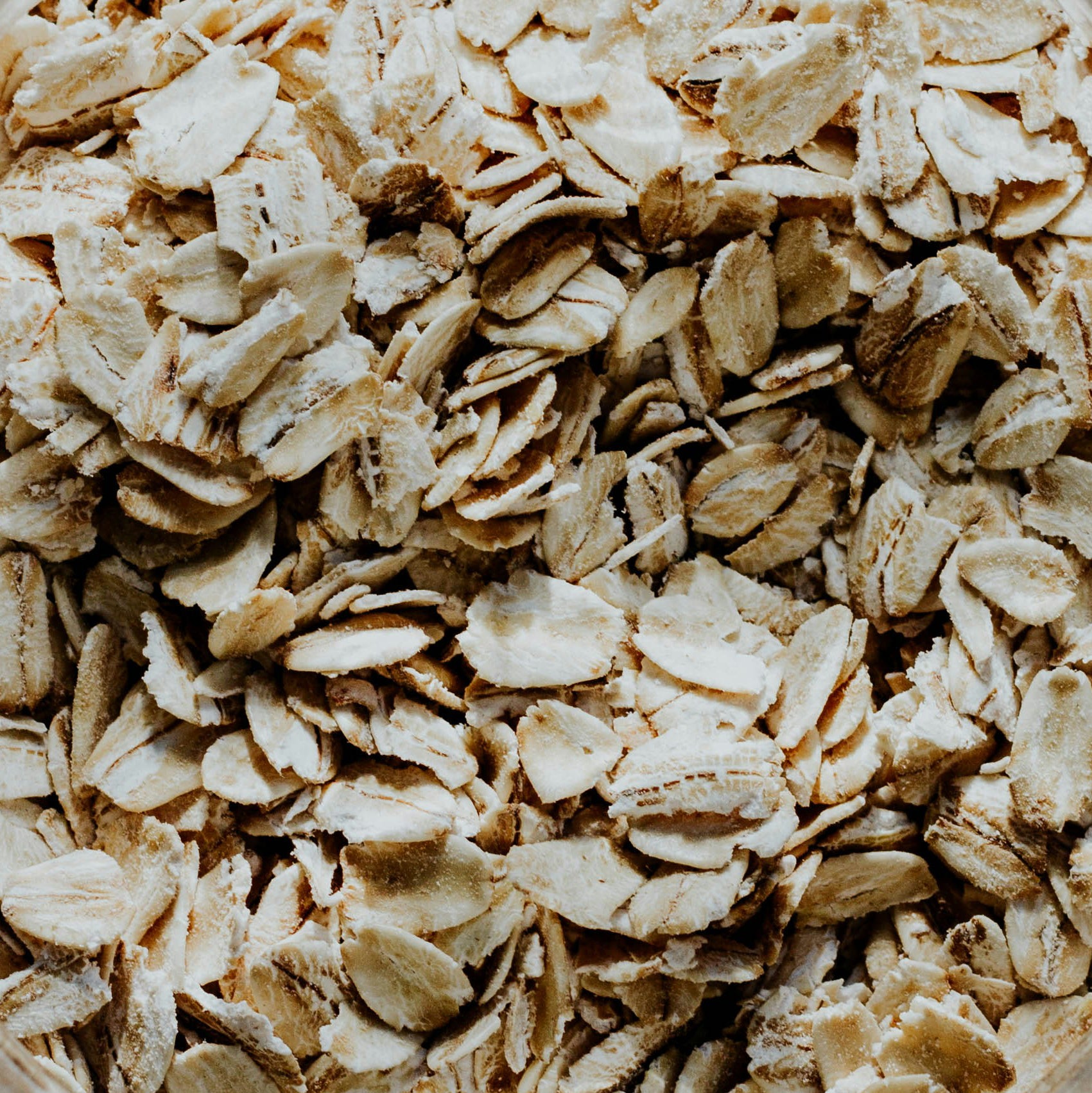 Close-up image of a pile of raw rolled oats. The oats are light brown and off-white, displaying their textured and slightly flattened surfaces. The image showcases the natural, unprocessed state of the oats, typically used for making oatmeal and other food products.