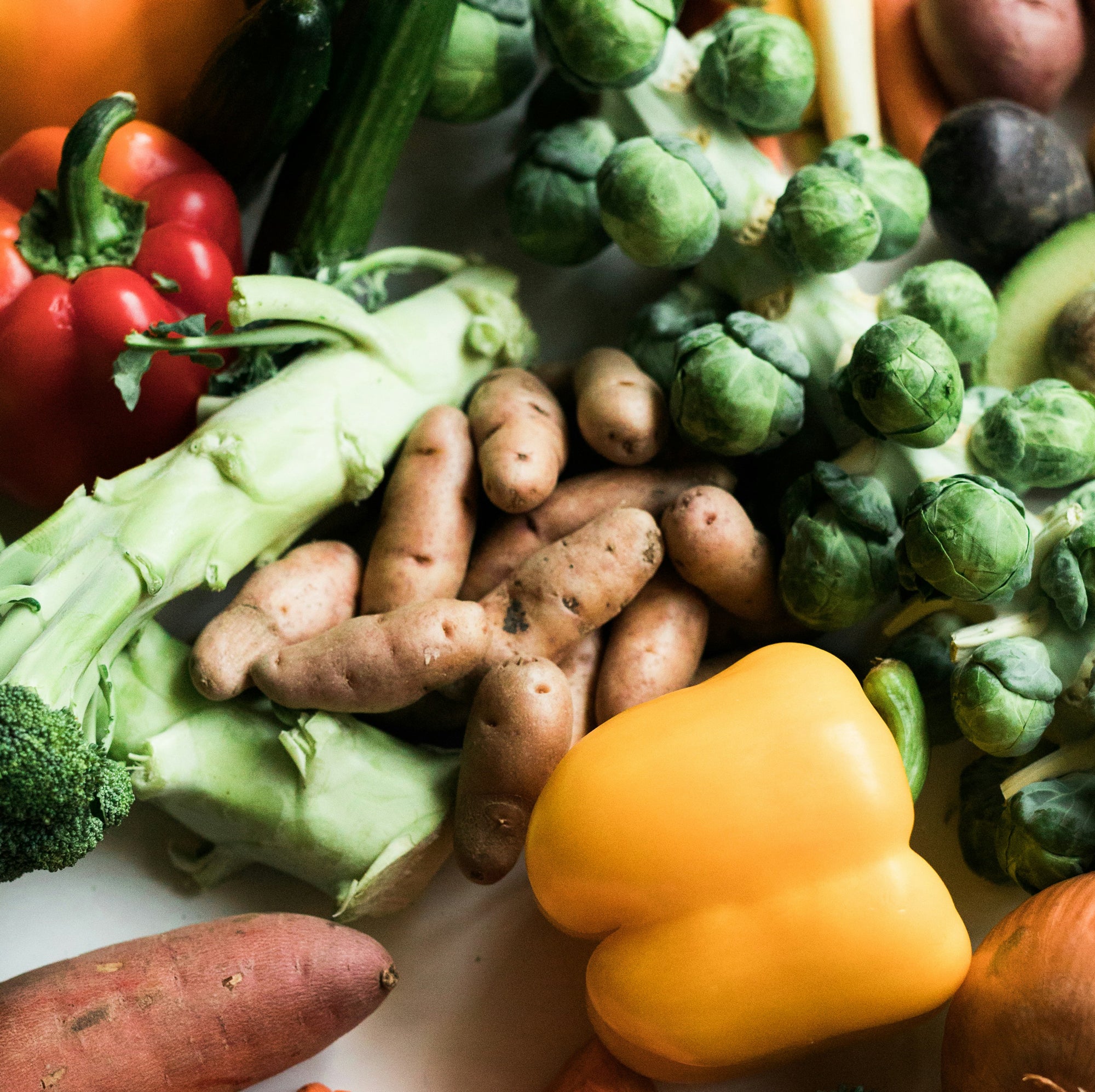 A variety of fresh vegetables are displayed, including yellow and red bell peppers, Brussels sprouts on a stalk, small potatoes, broccoli, cucumber, sweet potato, and some leafy greens. The vegetables are vibrant and arranged in a casual, overlapping manner.