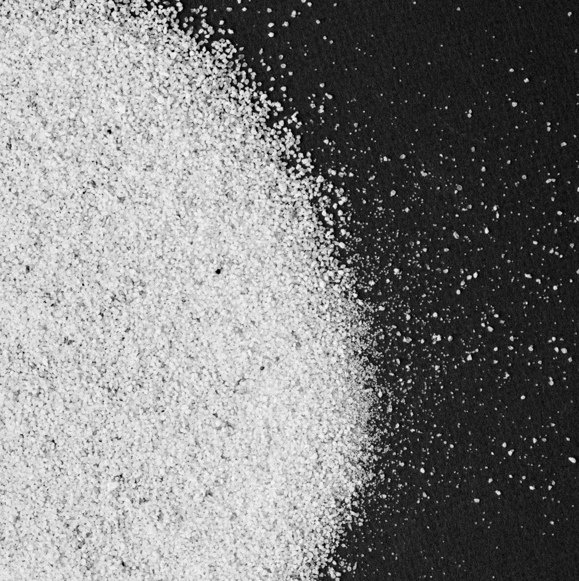 A close-up of white granules, resembling sugar or salt, scattered on a black surface. The granules are densely packed on the left side of the image and gradually disperse towards the right.