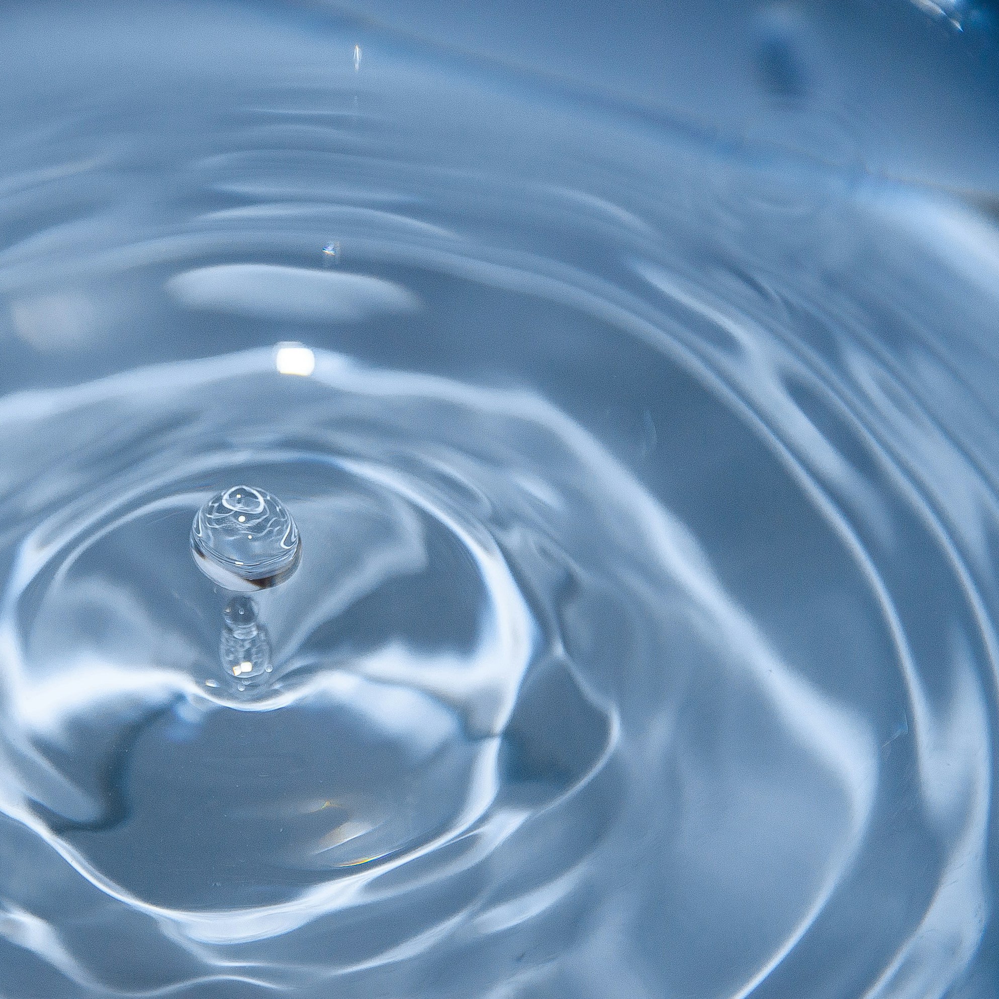 A close-up shot of a droplet of water creating ripples as it splashes into a calm water surface. The image captures the moment the droplet makes contact, forming concentric circles spreading outward in the clear blue water.